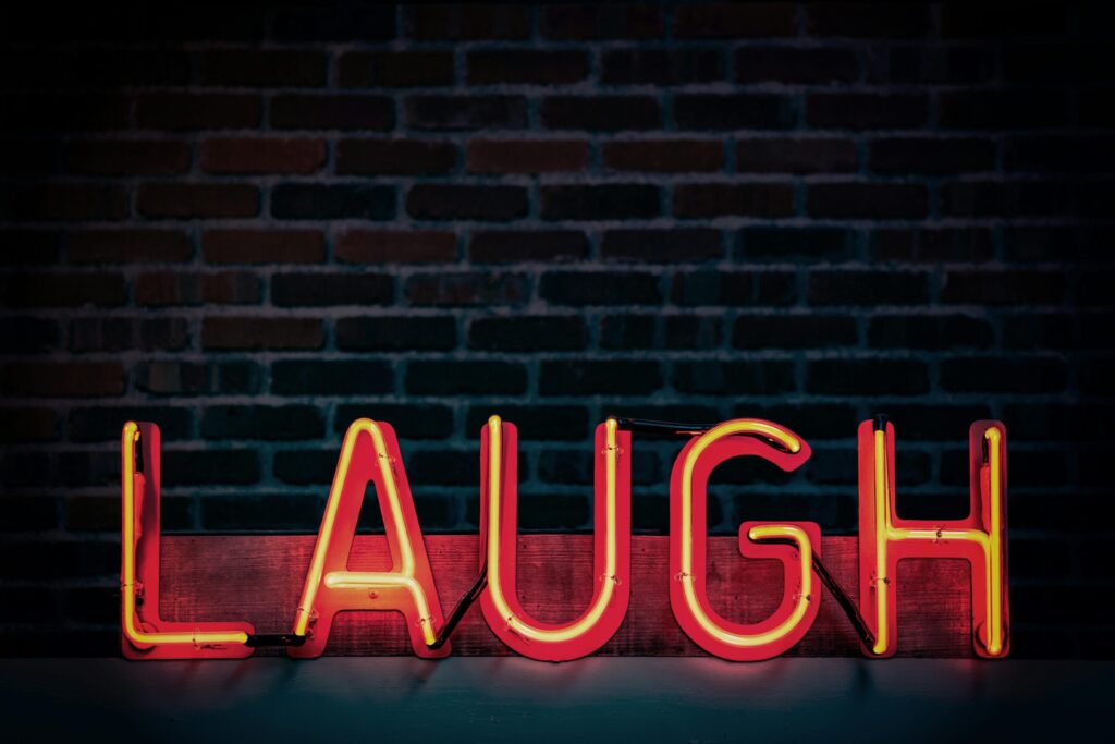 Wie sich Humor auf unsere Psychoe auswirkt: Auf dem Bild ist eine Leuchtschrift mit dem Schriftzug "Laugh" zu sehen, im Hintergrund eine dunkle Ziegelmauer