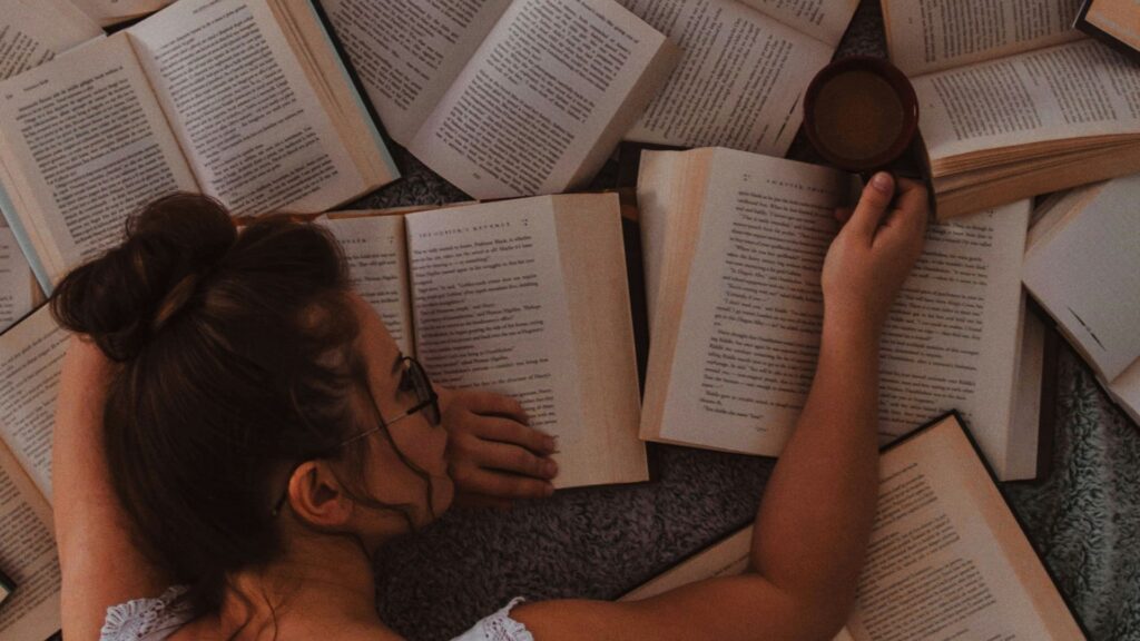 Frau liegt auf vielen offenen Bücher und hat einen Kaffe in der Hand.