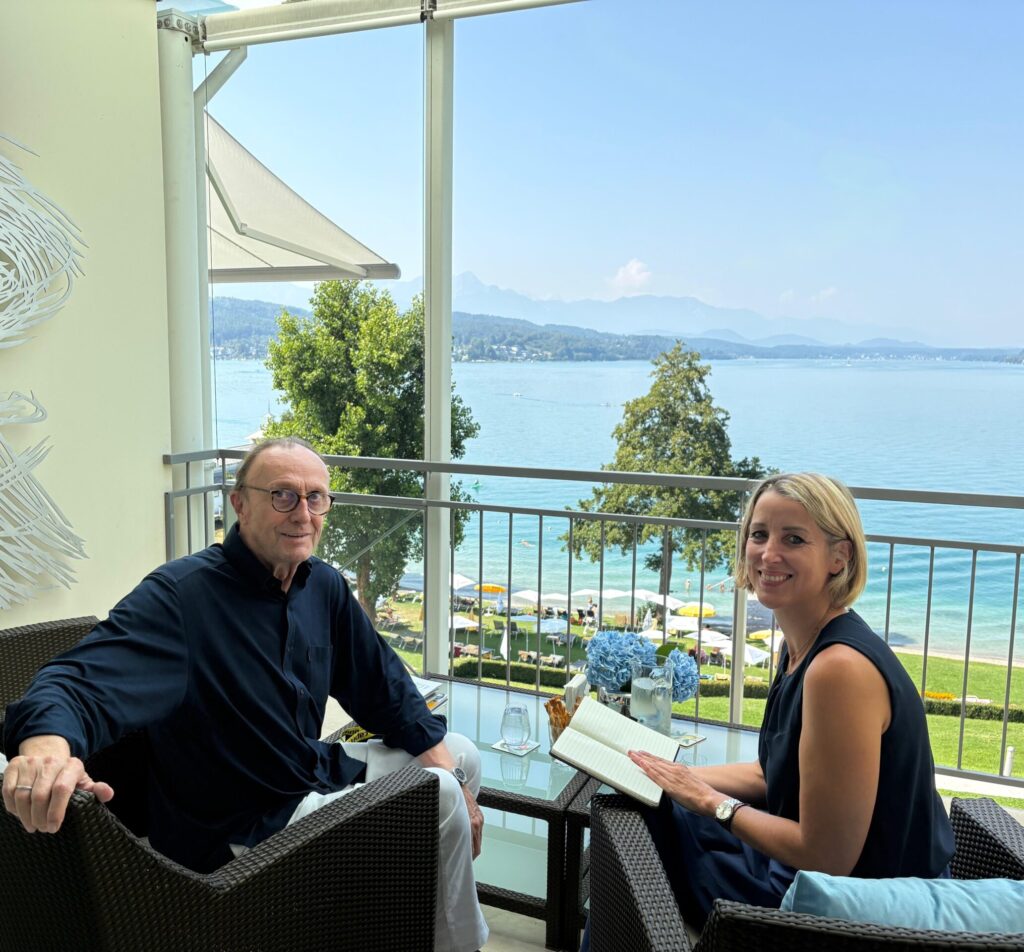 Ein Mann mit Brille und schwarzen Hemd und eine Frau mit blauem Shirt sitzen auf einer Terrasse vor dem Wörthersee