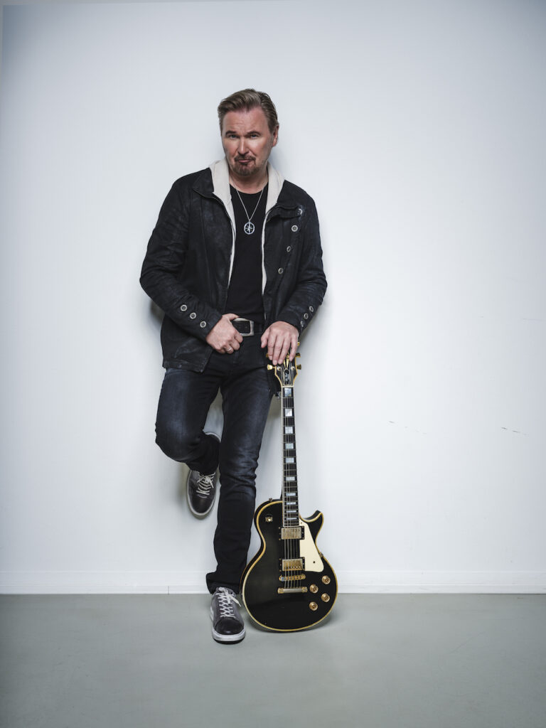 Der Körnern Sänger Nik P (ein Mann mit Bart und Lederjacke) lehnt an einer Wand und hält seine Gitarre