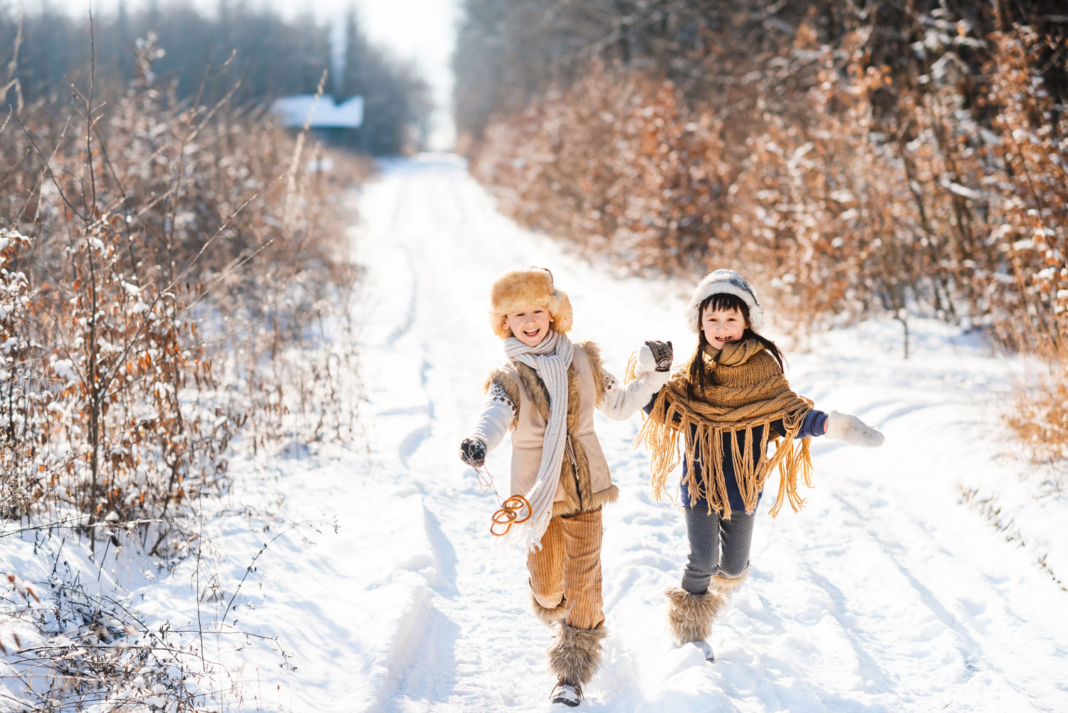Zwei Kinder laufen glücklich auf einem schneeigen Weg im Wald mit Haus im Hintergrund und blätterarmen Büschen an den Seiten.