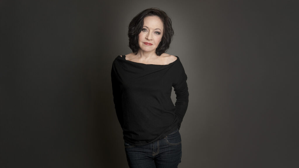 Maria Bill: Ganzkörperporträt einer Frau in schwarzem Oberteil vor neutralem Hintergrund.