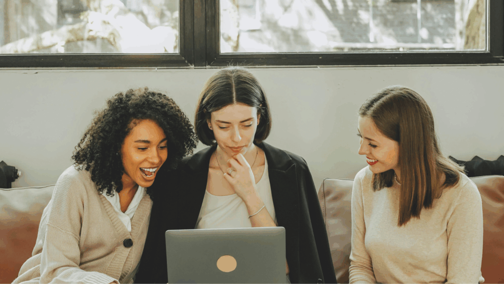 Drei Frauen sitzen gemeinsam auf einem Sofa. Die mittlere schaut auf ihren Laptop, die anderen lächeln.
