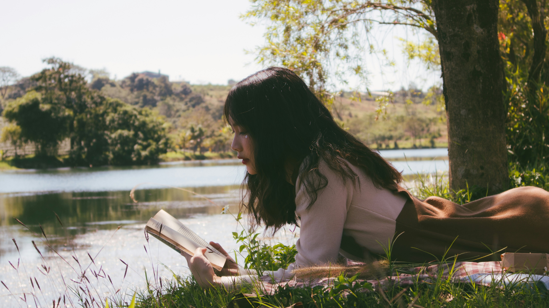 Eine Frau liegt auf dem Bauch und liest ein Buch. Im Hintergrund sind man einen See und Bäume