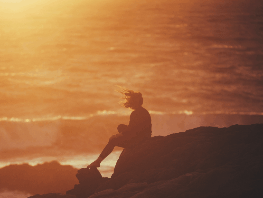 Eine Frau sitzt an einem Felsen bei Sonnenuntergang und schaut auf das Meer